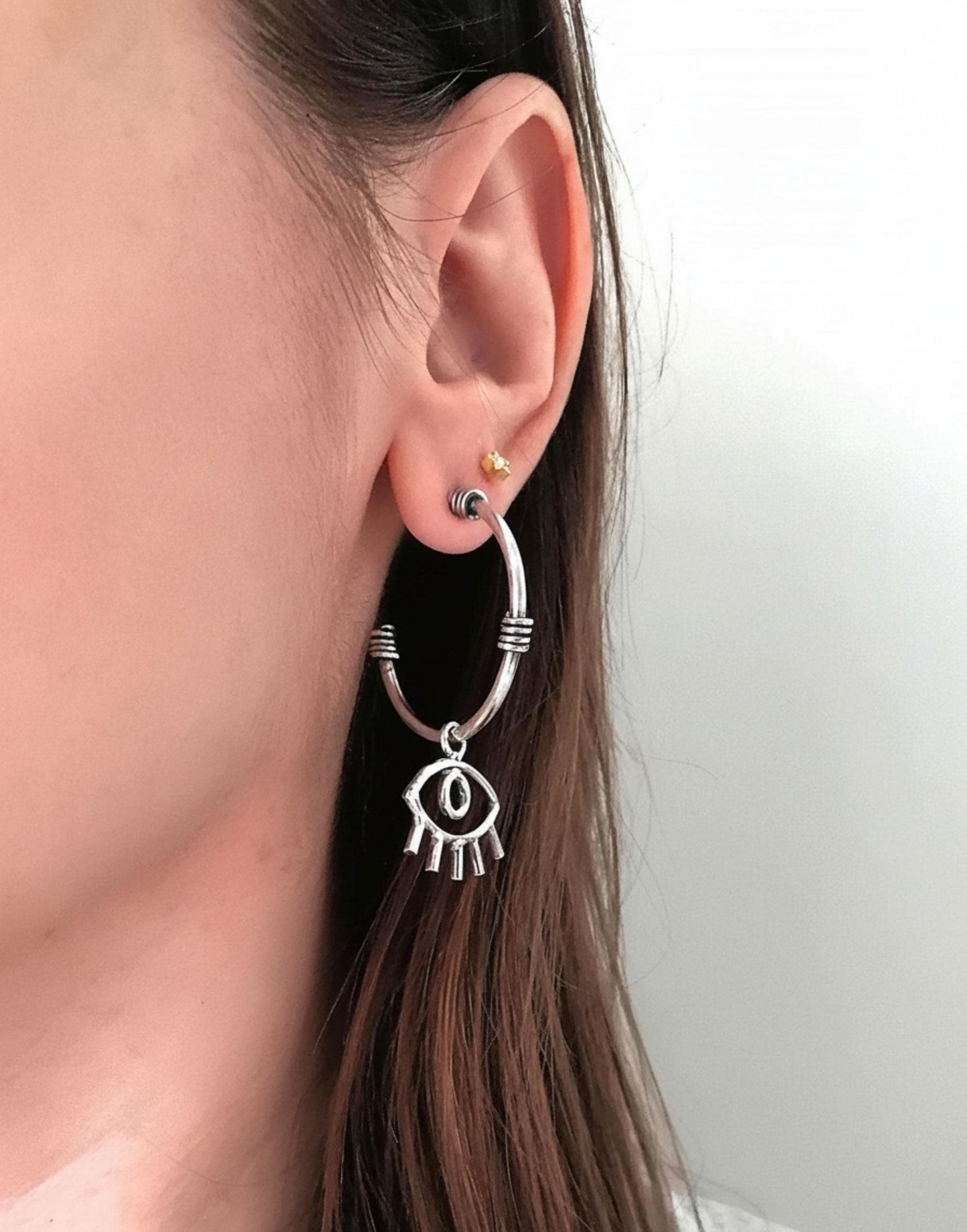 Close-up of ear wearing silver hoop earring with eye-shaped charm and small gold stud
