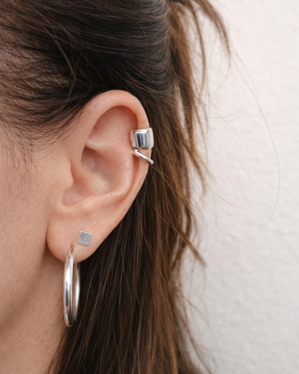 Close-up of an ear with silver hoop earrings and a small stud, against a neutral background.