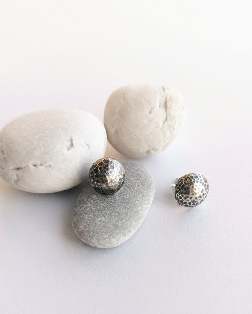 Two small silver-toned textured stud earrings displayed on smooth gray stones against a clean white background.