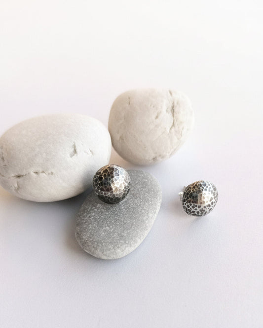 Two small silver-toned textured stud earrings displayed on smooth gray stones against a clean white background.