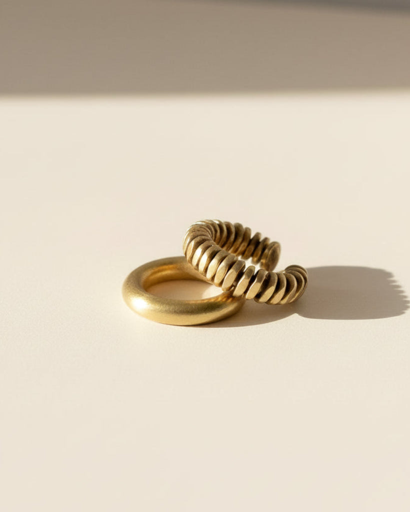 Two gold rings on a warm cream surface under direct sunlight, with one smooth band ring and one textured, ribbed spiral ring.