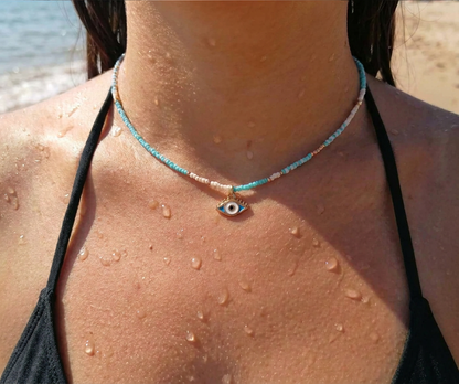 A close-up of a person at the beach wearing a delicate beaded choker necklace with a small evil eye pendant. The necklace features light blue, pink, and white beads with a silver-toned evil eye charm in the center.