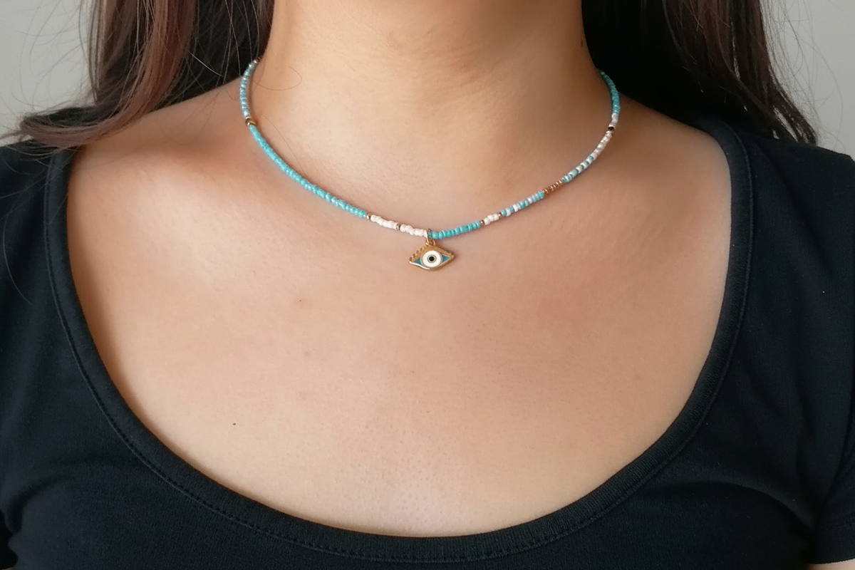 A close-up of a person's neck and chest wearing a delicate beaded necklace with a small gold evil eye pendant.