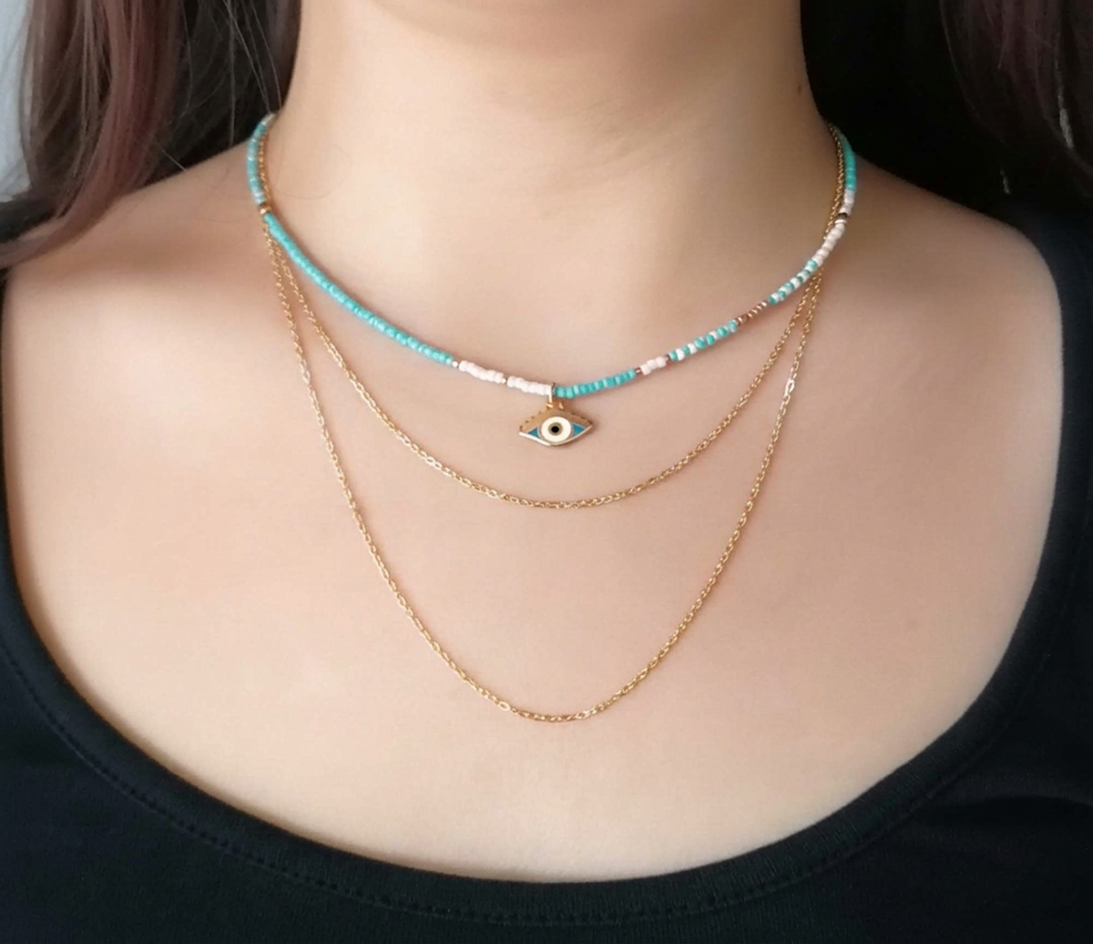 A close-up of a person's neck and chest wearing a layered necklace set. The set includes a delicate gold chain, a longer gold chain, and a turquoise beaded choker necklace with a central gold evil eye charm.