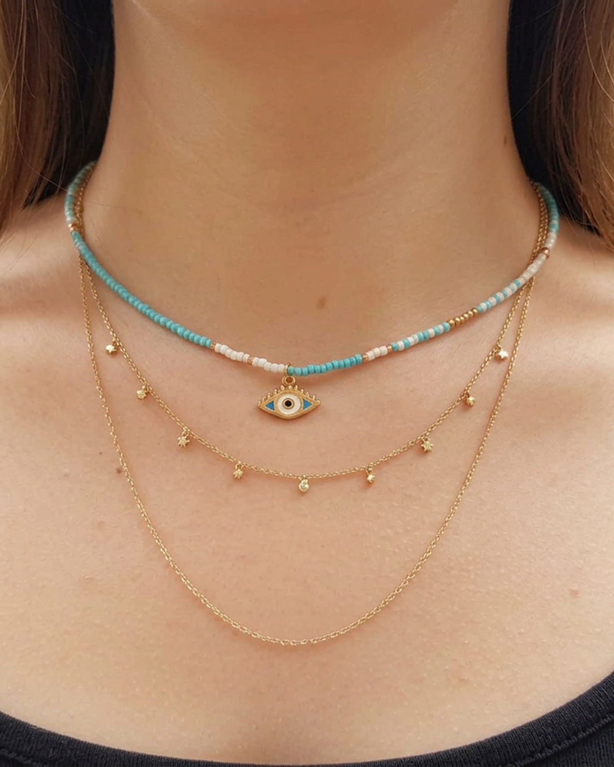 A close-up of a person's neck and upper chest wearing a layered necklace set. The set includes a turquoise and white beaded choker with a gold evil eye pendant, a gold chain with small gold beads, and a longer gold chain.