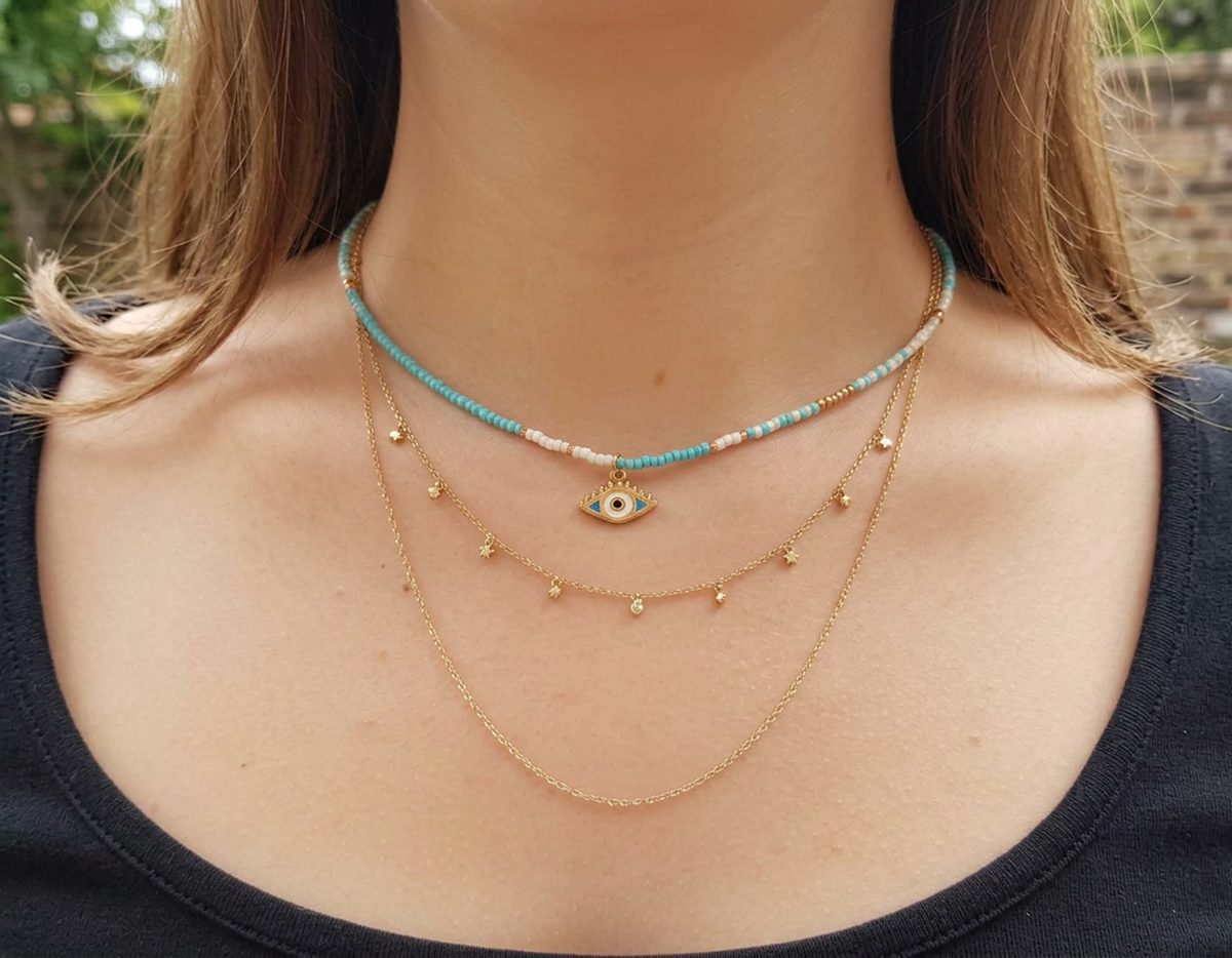 A close-up of a person's neck and upper chest wearing a layered necklace set. The set includes a turquoise and white beaded choker with a gold evil eye pendant, a gold chain with small gold beads, and a longer gold chain.