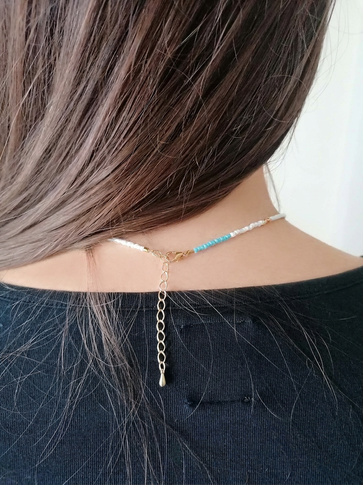 A close-up back view of a person with long brown hair wearing a delicate choker necklace. The necklace features a gold chain, a small section of light blue beads, and clear rhinestone-like accents.