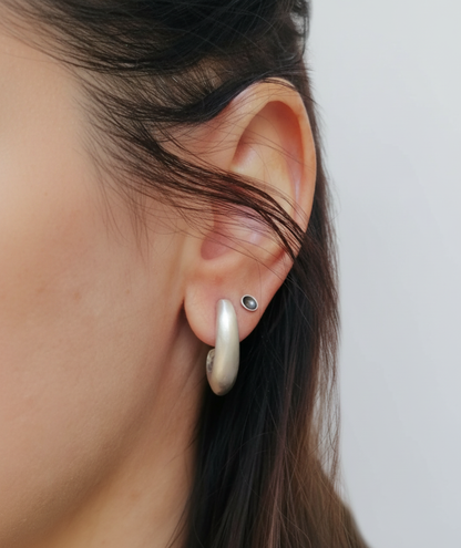 A close-up of a person's ear wearing a silver hoop earring and a small silver stud with a dark gemstone, against a plain light gray background.