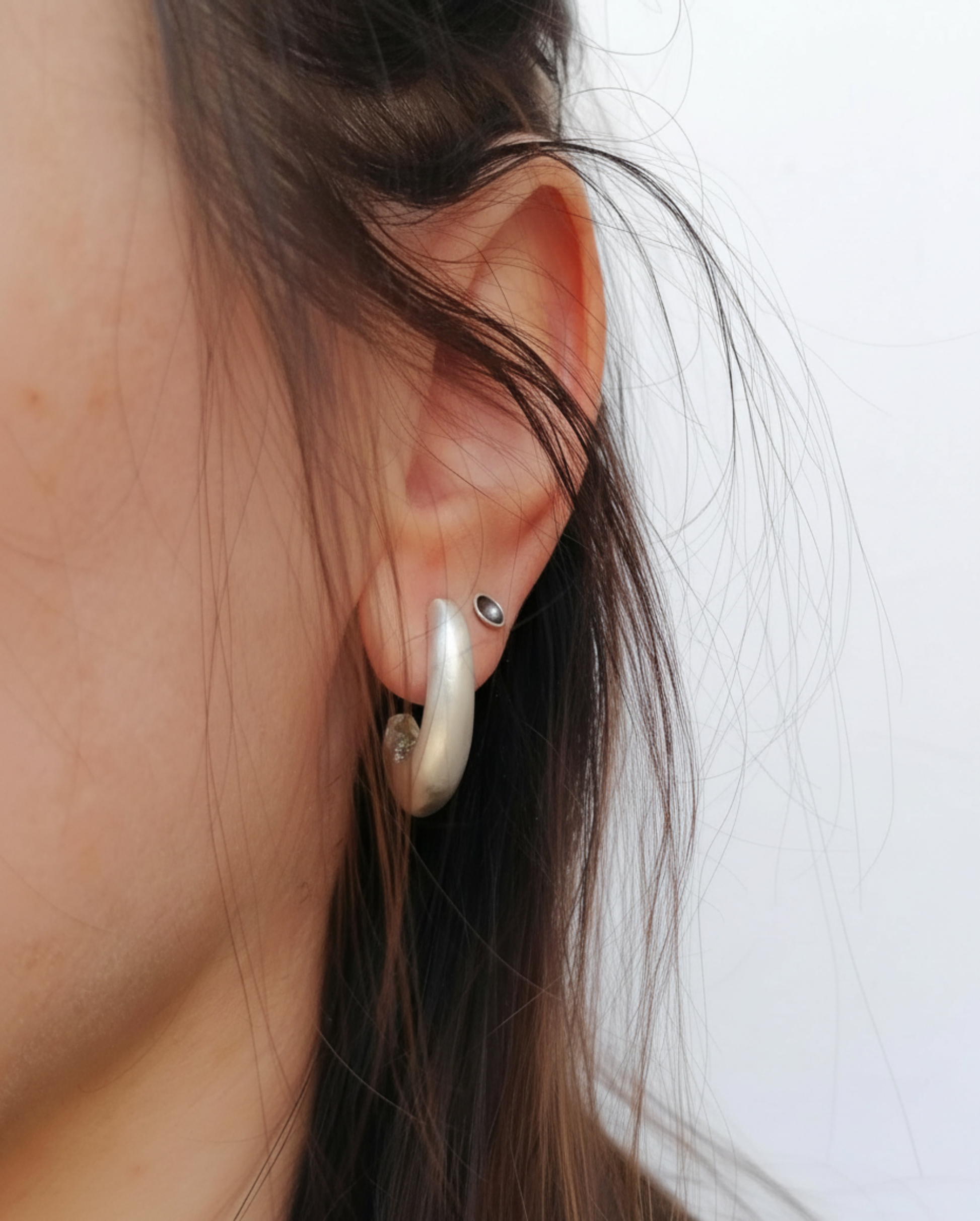 A close-up of a person's ear wearing a large, curved silver hoop earring and a small silver stud earring against a plain light background.