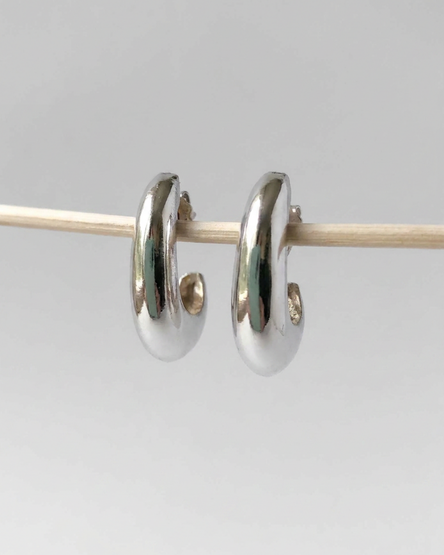 A pair of polished silver huggie hoop earrings hanging from a thin wooden rod against a light gray background.