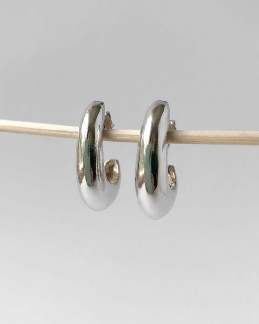 A pair of polished silver huggie hoop earrings hanging from a thin wooden rod against a light gray background.