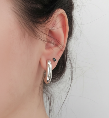 A close-up of a person's ear wearing a polished silver hoop earring and a small black stud earring against a light gray background.