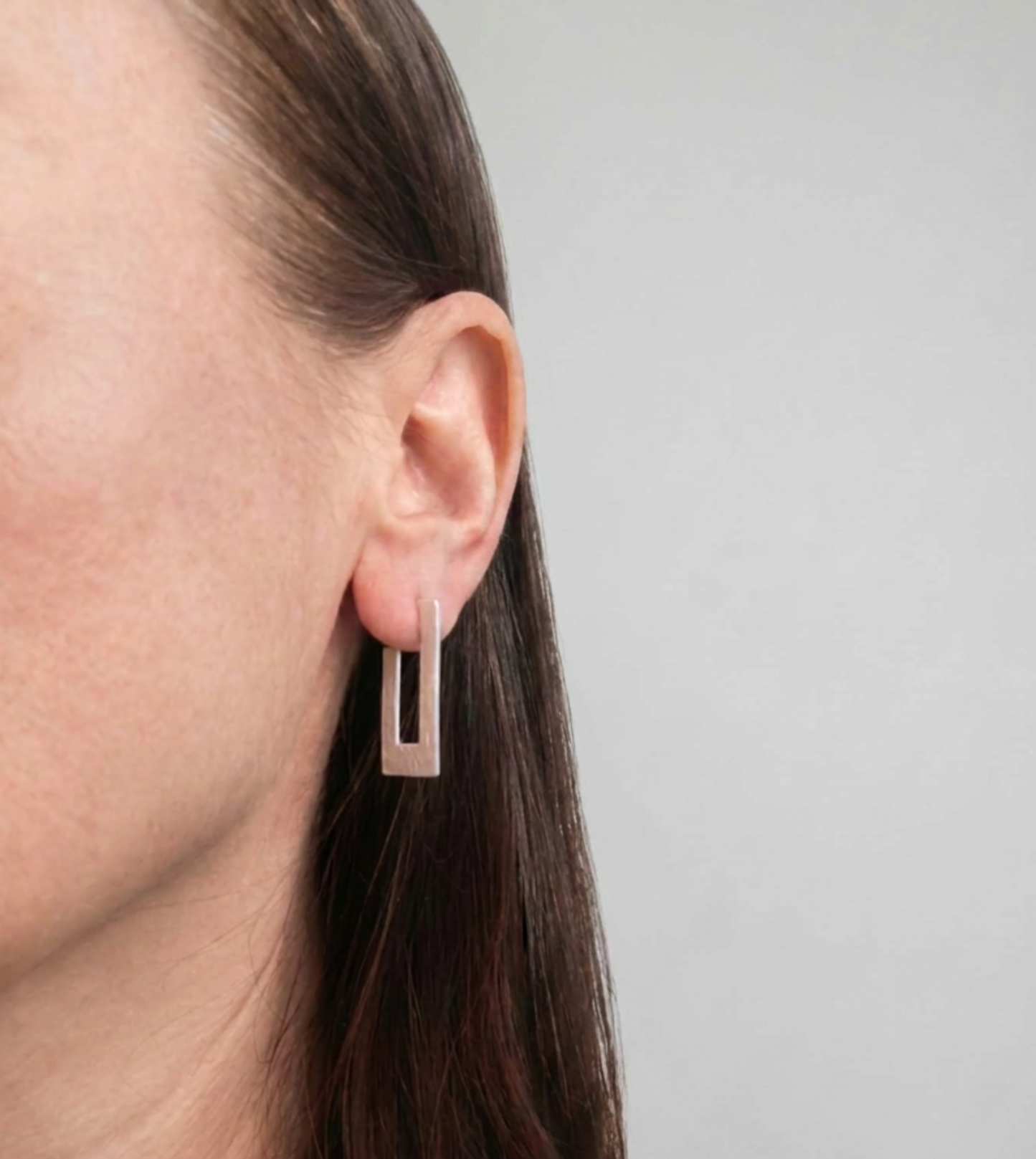 A close-up of a woman's ear wearing a minimalist, rectangular rose gold hoop earring against a plain light gray background.