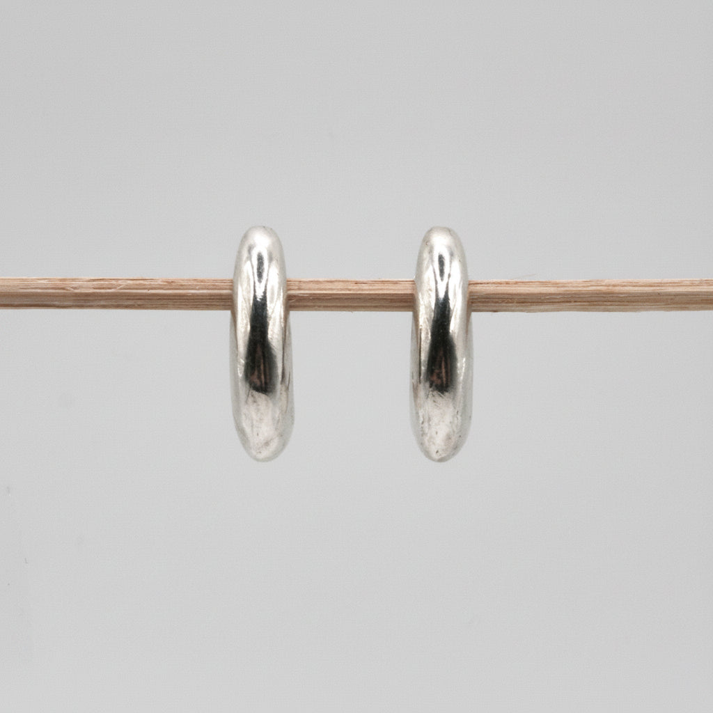 A pair of polished silver hoop earrings hanging from a thin wooden stick against a plain light gray background.