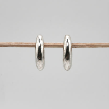 A pair of polished silver hoop earrings hanging from a thin wooden stick against a plain light gray background.