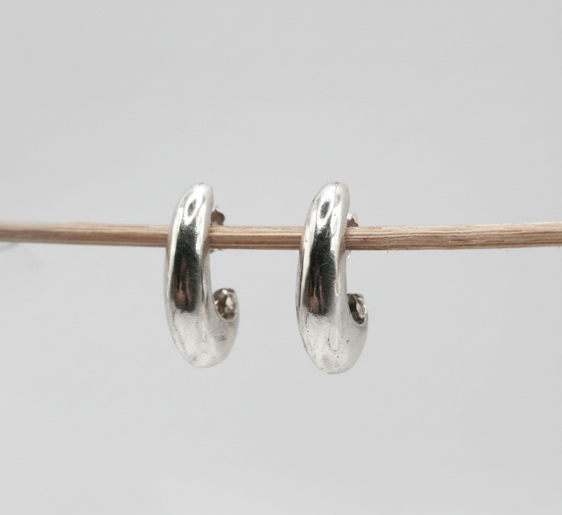 A pair of polished silver huggie hoop earrings hanging from a thin wooden stick against a plain light gray background.