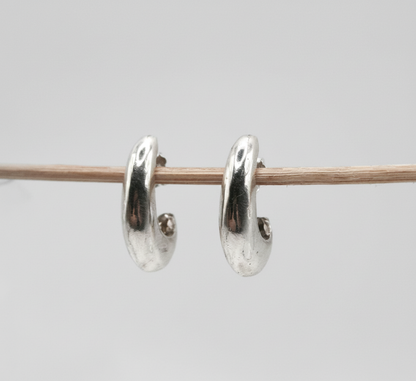 A pair of polished silver huggie hoop earrings hanging from a thin wooden stick against a plain light gray background.