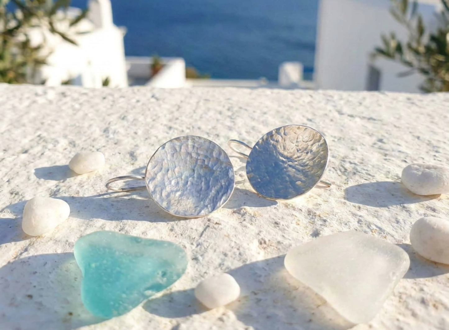 A pair of hammered silver disc earrings resting on a white stone surface outdoors, surrounded by small white stones and a piece of blue sea glass, with a blurred coastal background.