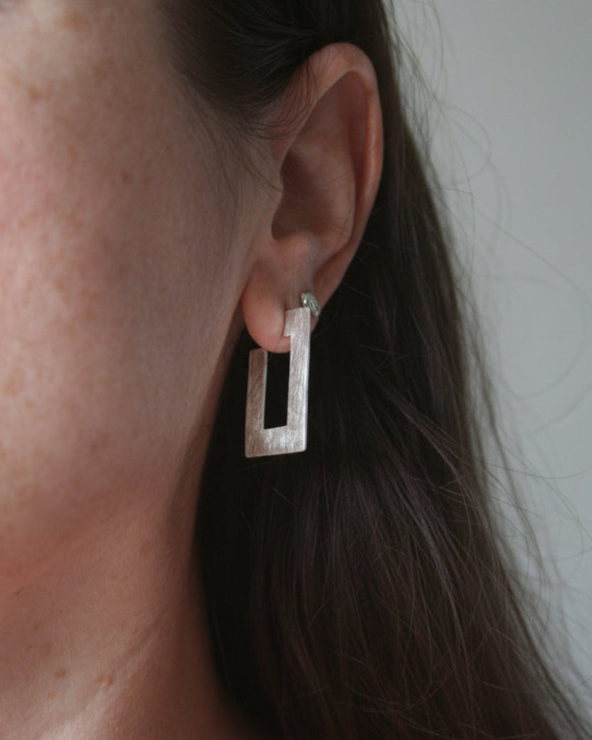 A close-up of a person's ear wearing a textured silver geometric rectangular stud earring.