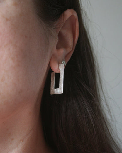 A close-up of a person's ear wearing a textured silver geometric rectangular stud earring.