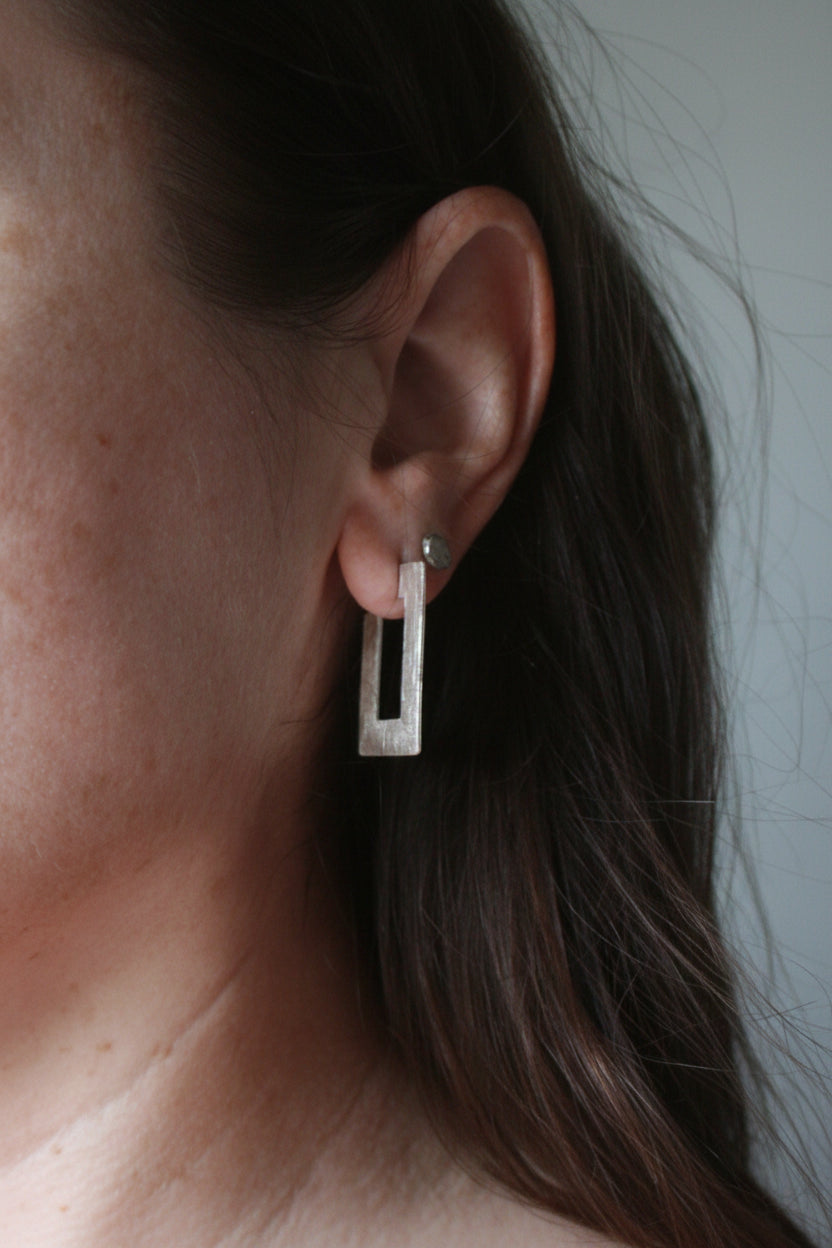 A close-up of a person's ear wearing a modern geometric silver dangle earring with a rectangular frame design.