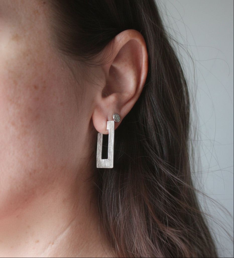 A close-up shot of a woman's ear wearing a small silver rectangular stud earring with a black rectangular inlay.