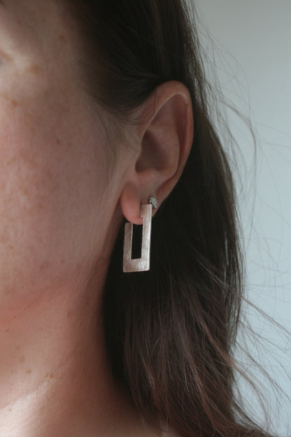 A close-up of a person's ear wearing a geometric silver drop earring with a rectangular cutout design.