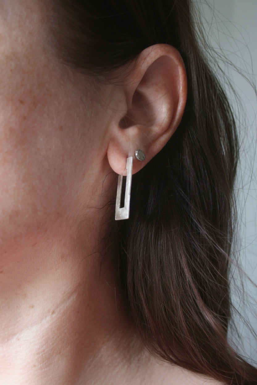 A close-up of a person's ear wearing a modern silver geometric drop earring with a vertical rectangular bar and a small round stud earring.