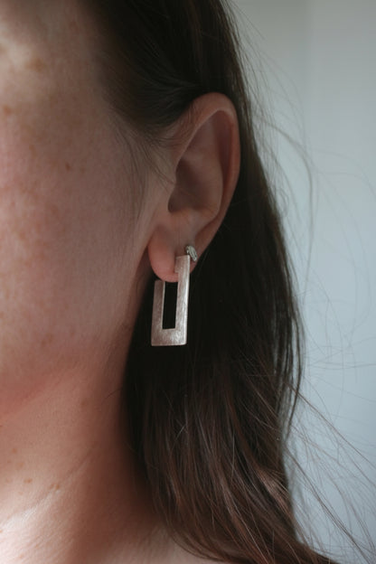 A close-up shot of a woman's ear wearing a small silver-toned rectangular stud earring.