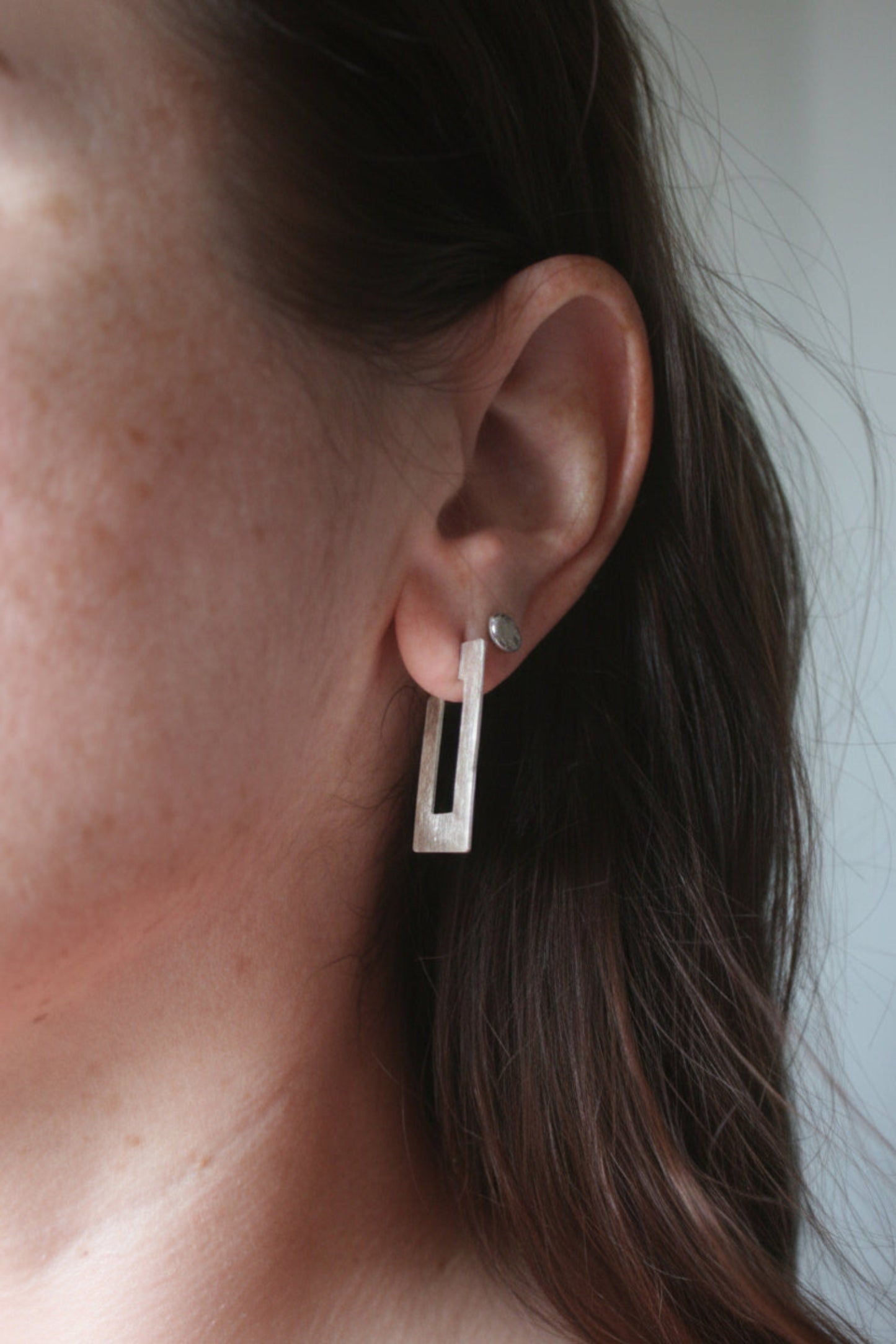 A close-up of a person's ear wearing a modern geometric silver dangle earring with a vertical rectangular frame and a small round stud earring behind it.