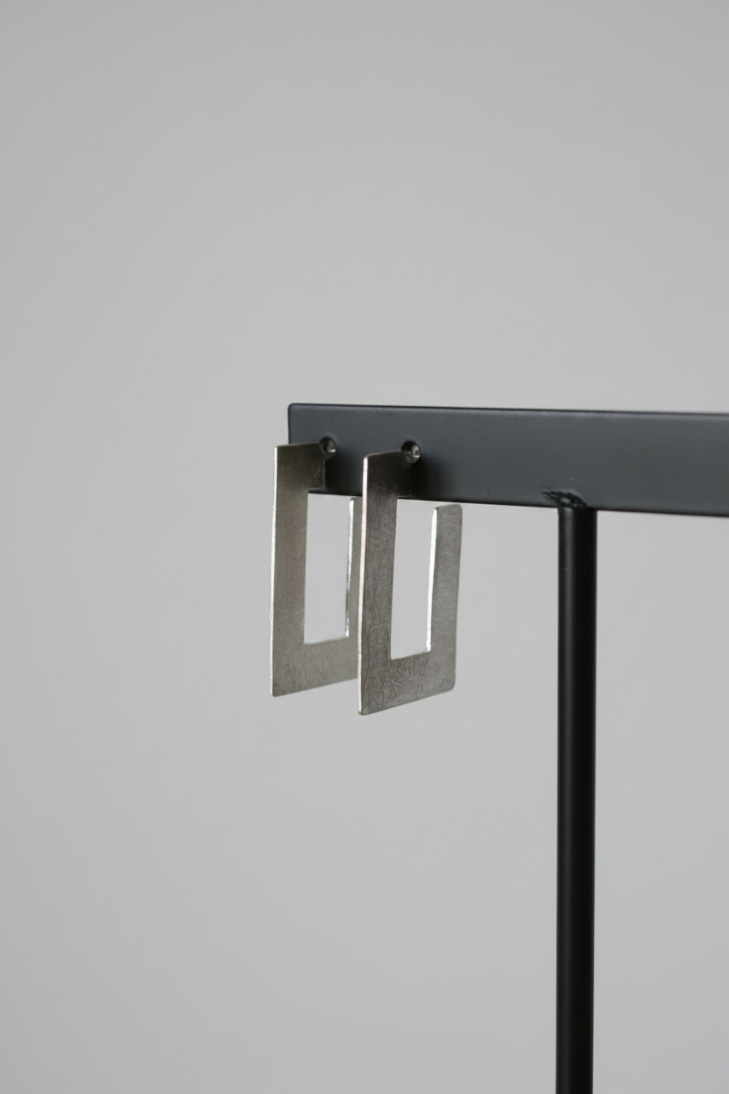 A pair of silver-toned geometric square drop earrings hanging from a black display stand against a textured light gray wall.