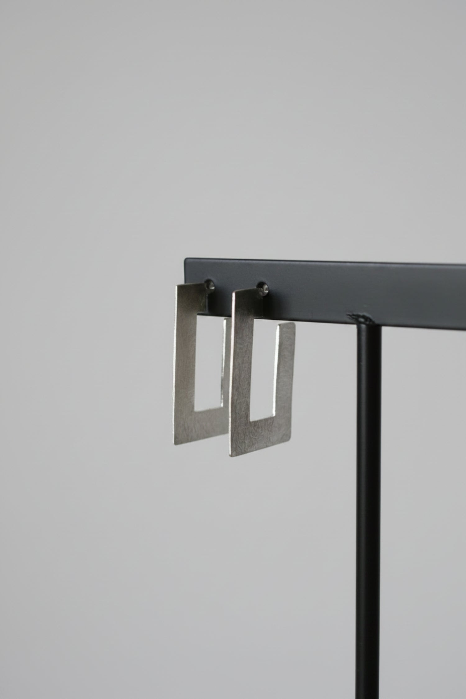 A pair of silver-toned geometric square drop earrings hanging from a black display stand against a textured light gray wall.