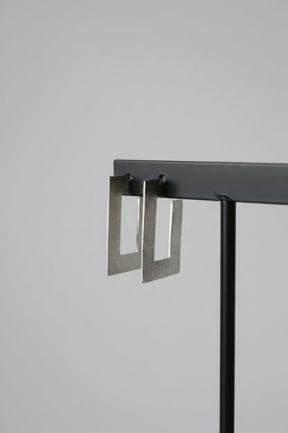 A pair of silver-toned geometric square drop earrings hanging from a black display stand against a textured light gray wall.