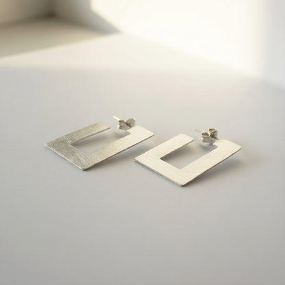 A pair of textured silver geometric dangle earrings with a square open-center design, resting on a plain gray surface.