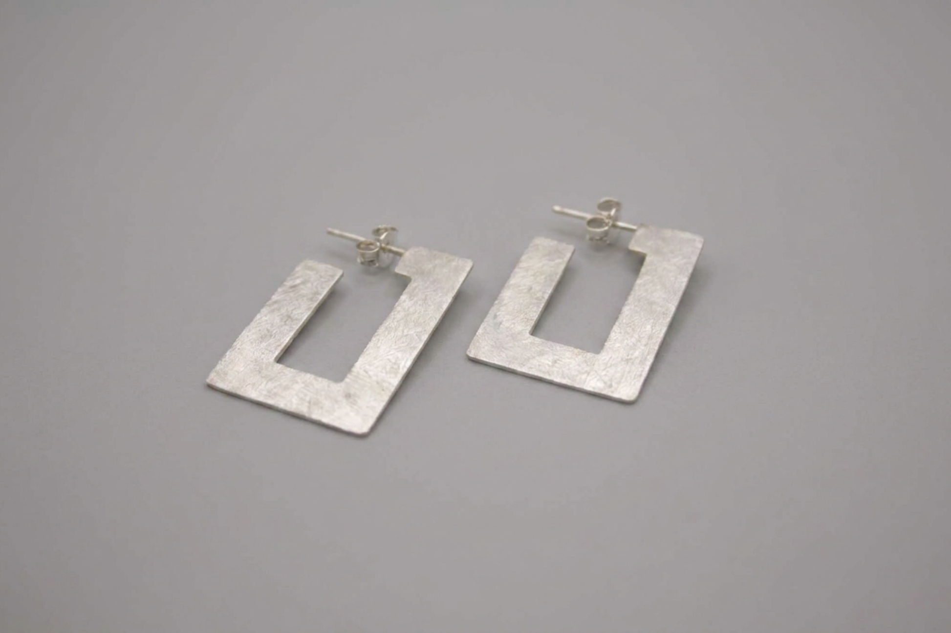 A pair of textured silver geometric drop earrings with a square cutout design, displayed on a neutral gray background.