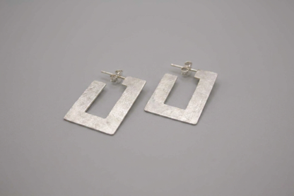 A pair of textured silver geometric drop earrings with a square cutout design, displayed on a neutral gray background.