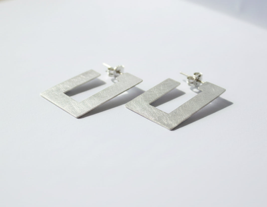 A pair of textured silver geometric drop earrings with a square cutout design, resting on a light gray surface.