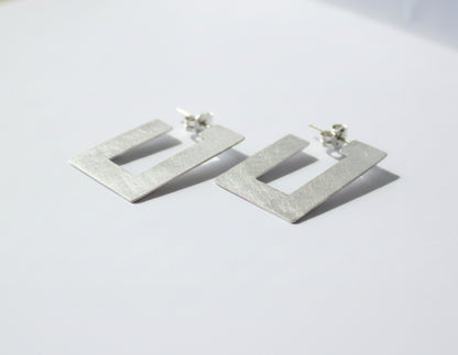 A pair of textured silver geometric drop earrings with a square cutout design, resting on a light gray surface.