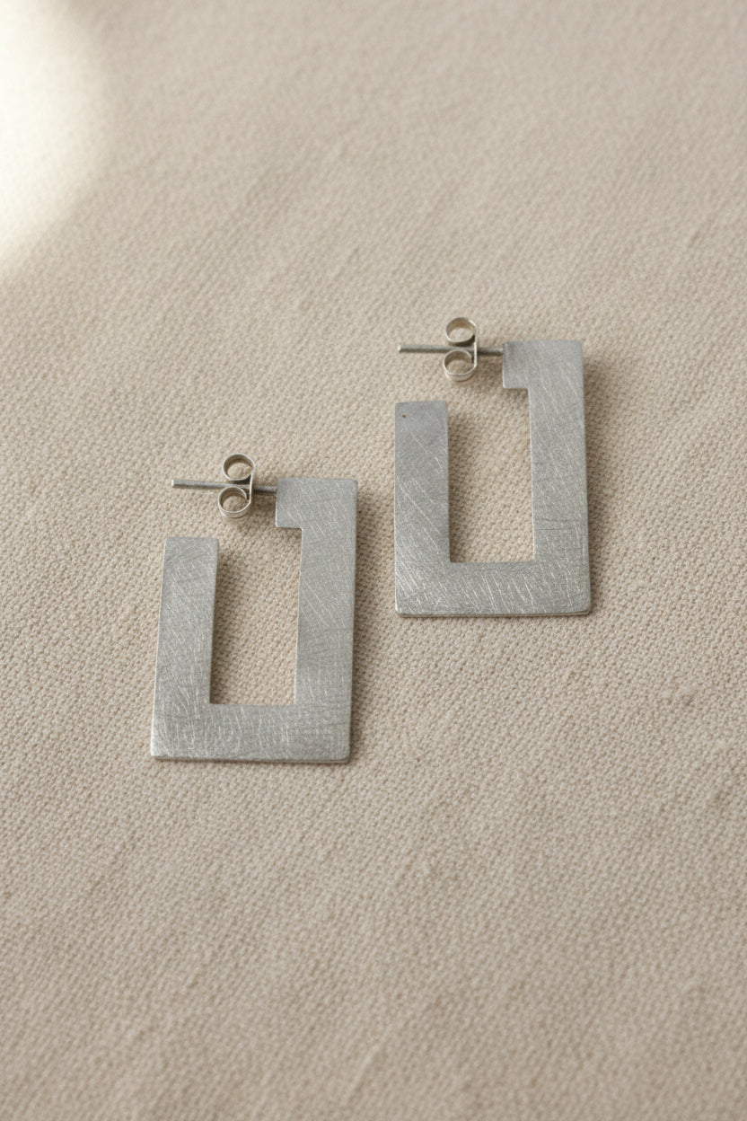 A pair of textured silver geometric drop earrings with open rectangular shapes, displayed on a plain gray background.