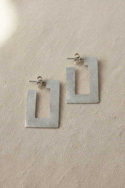 A pair of textured silver geometric drop earrings with open rectangular shapes, displayed on a plain gray background.