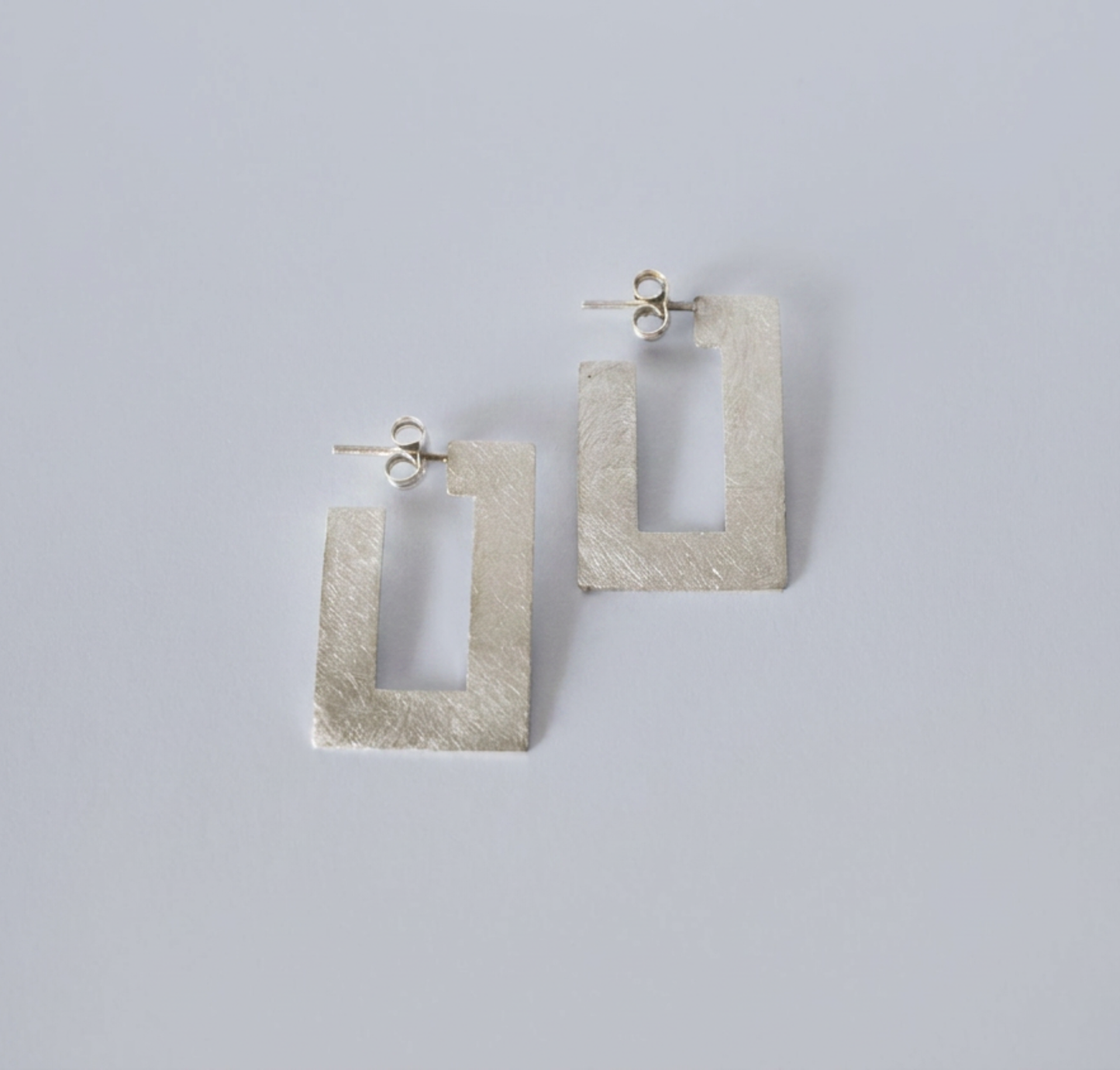 A pair of textured silver geometric drop earrings with a rectangular open-center design, displayed on a clean light gray background.