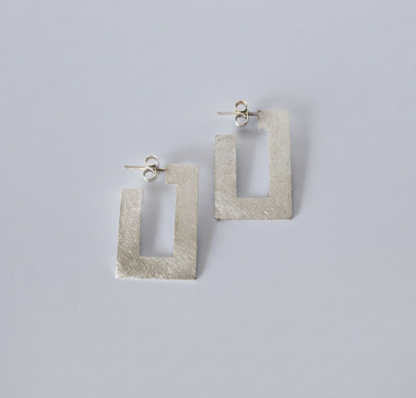 A pair of textured silver geometric drop earrings with a rectangular open-center design, displayed on a clean light gray background.