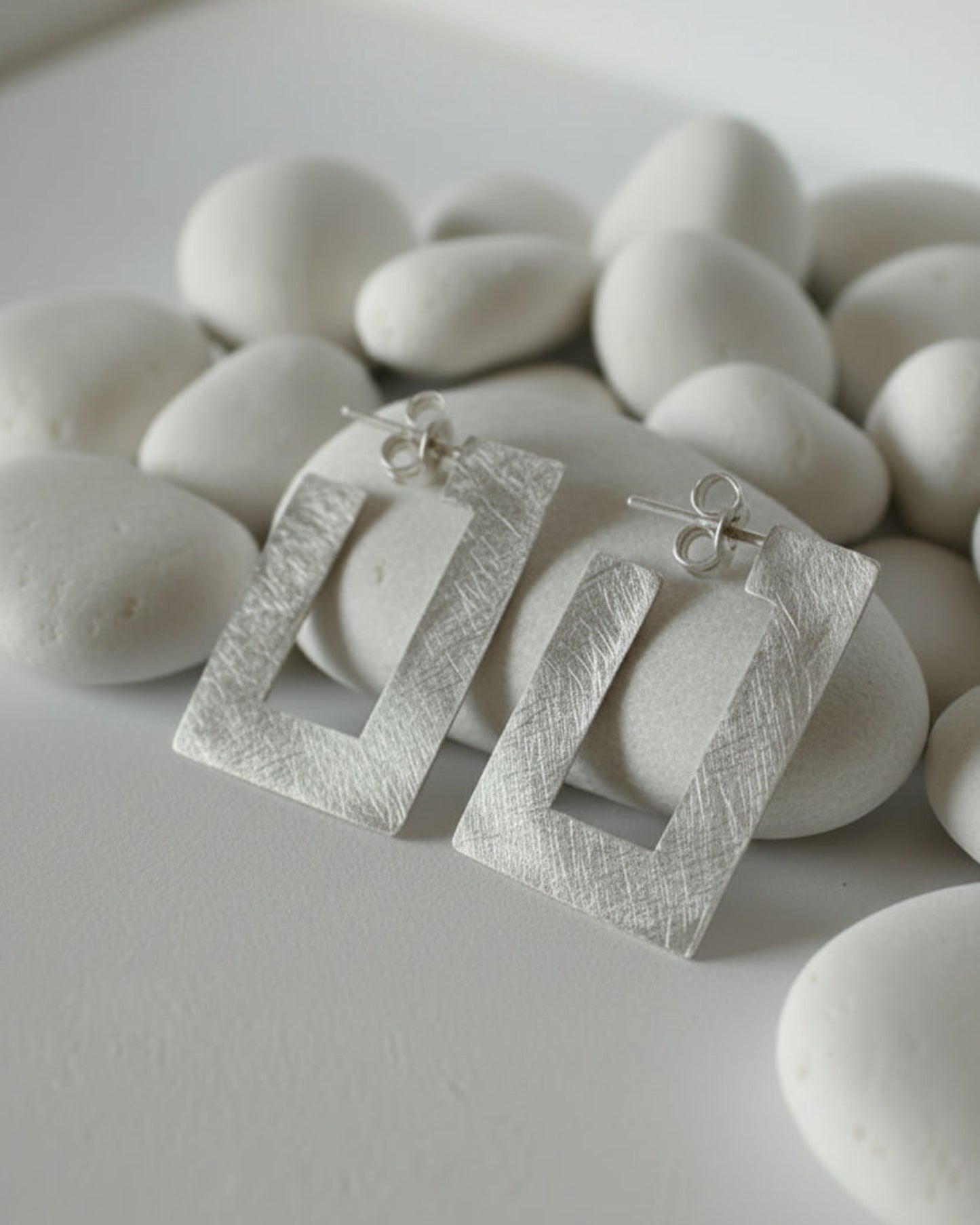A pair of textured silver geometric dangle earrings with open rectangular frames, displayed on a small white shell stand against a neutral gray background.