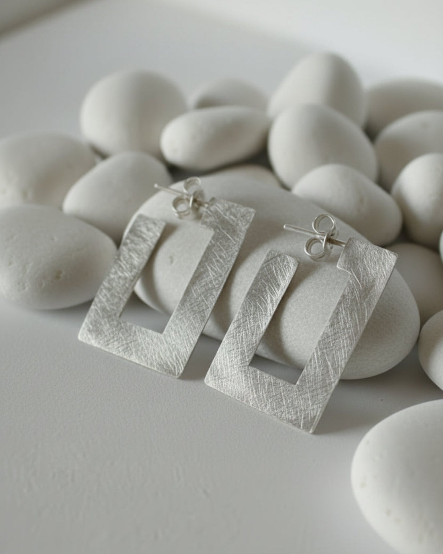 A pair of textured silver geometric dangle earrings with open rectangular frames, displayed on a small white shell stand against a neutral gray background.