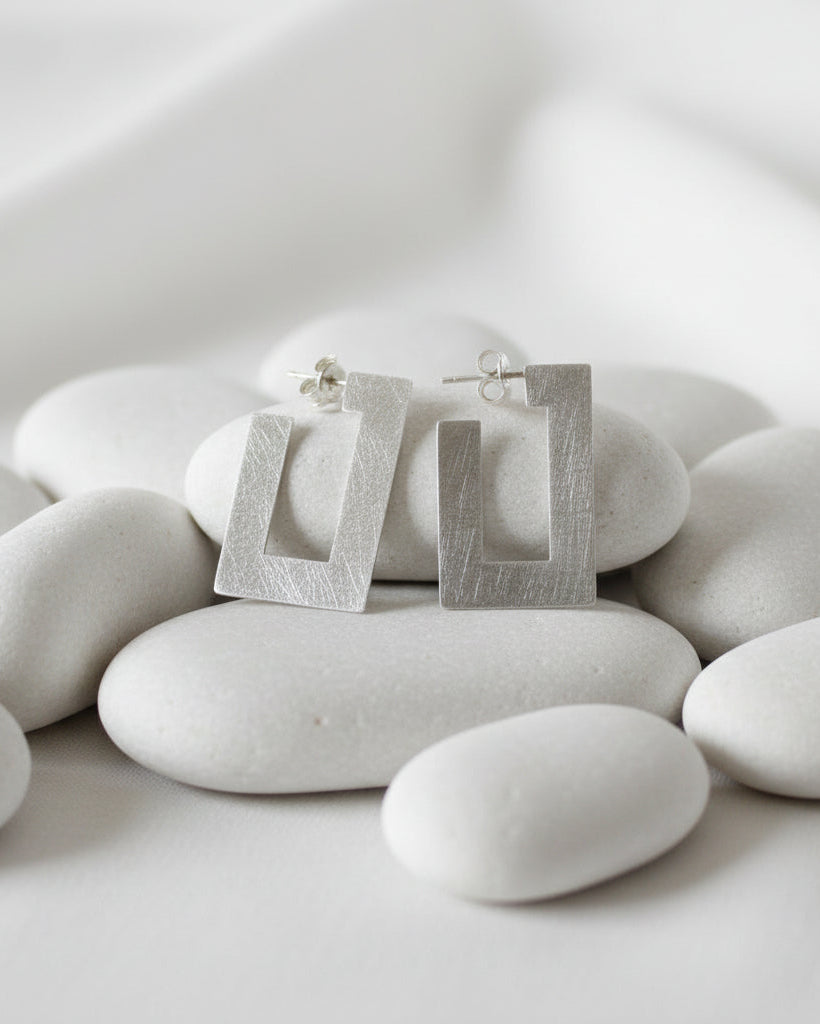 A pair of textured silver geometric stud earrings with a square frame design, displayed on smooth white stones against a soft white background.