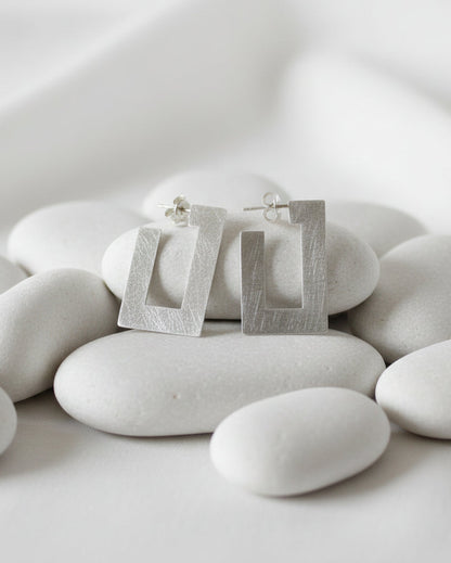 A pair of textured silver geometric stud earrings with a square frame design, displayed on smooth white stones against a soft white background.