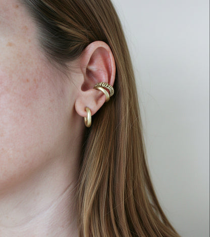 A close-up of a person's ear wearing a stack of gold earrings, including a small hoop and a textured ear cuff, against a plain light background.