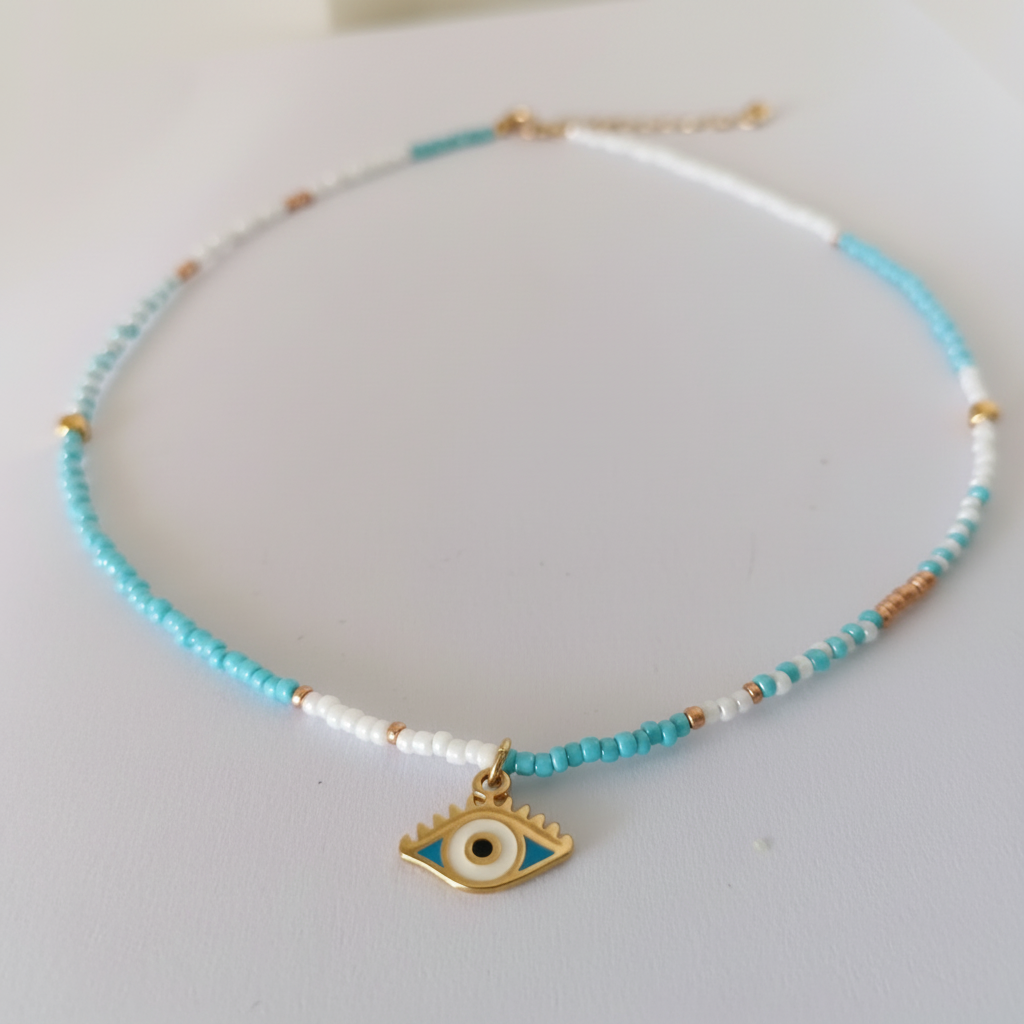 A delicate beaded necklace with turquoise, white, and rose gold beads, featuring a gold-tone evil eye charm at the center, displayed on a light off-white background.