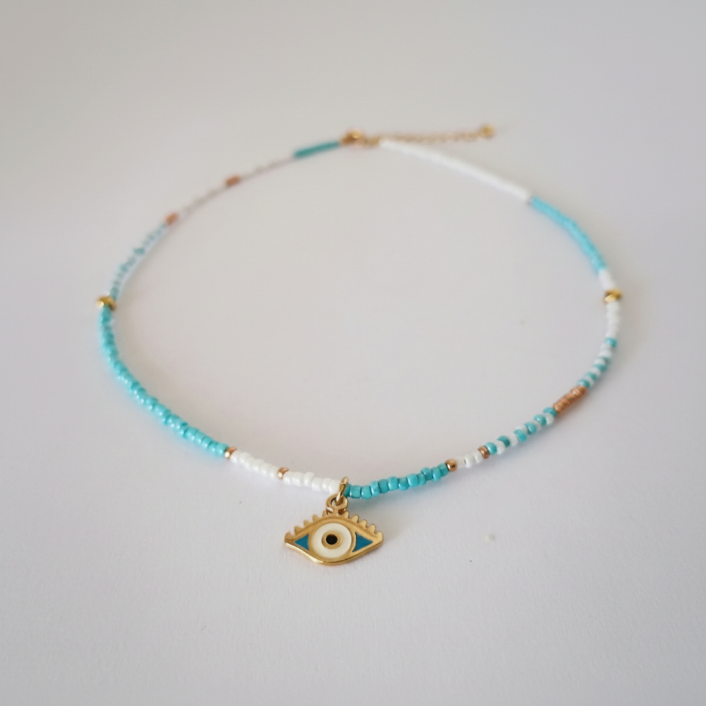 A delicate beaded necklace with light blue, white, and gold beads, featuring a gold-tone evil eye charm at the center, displayed on a light gray background.