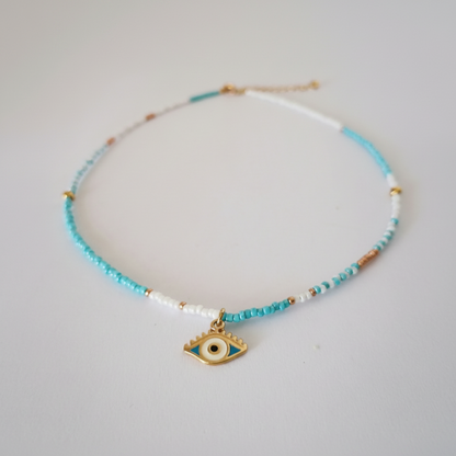 A delicate beaded necklace with light blue, white, and gold beads, featuring a gold-tone evil eye charm at the center, displayed on a light gray background.